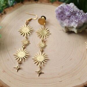 May include: A pair of gold-tone dangle earrings featuring multiple starburst shapes and star charms. The earrings are displayed on a wooden surface with a purple amethyst crystal.