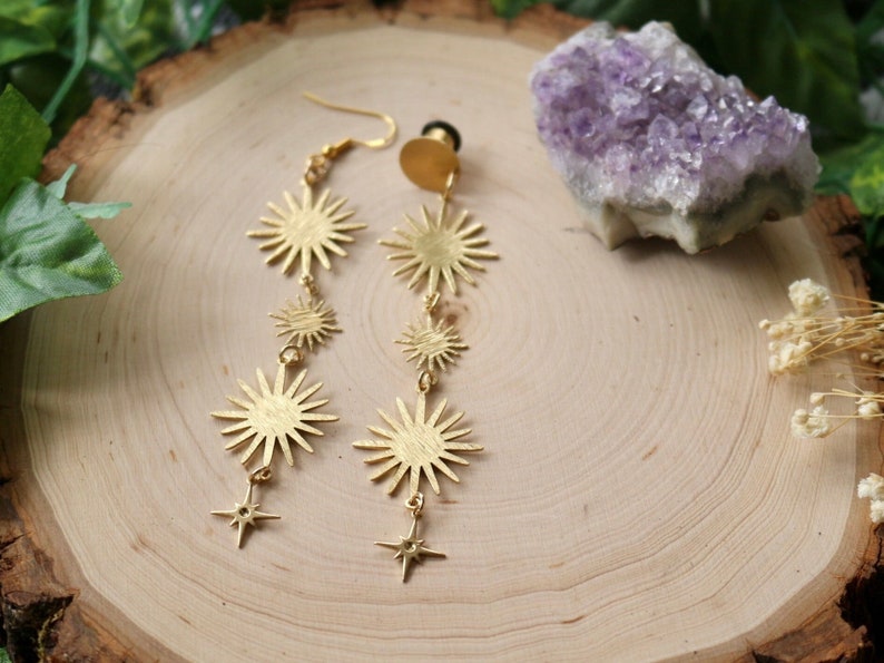 May include: A pair of gold-tone dangle earrings featuring multiple starburst shapes and star charms. The earrings are displayed on a wooden surface with a purple amethyst crystal.