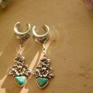 May include: A pair of silver-toned ear gauges with floral and turquoise heart-shaped pendants. The gauges are displayed on a light brown wooden surface, with a sprig of lavender and a purple crystal in the background.