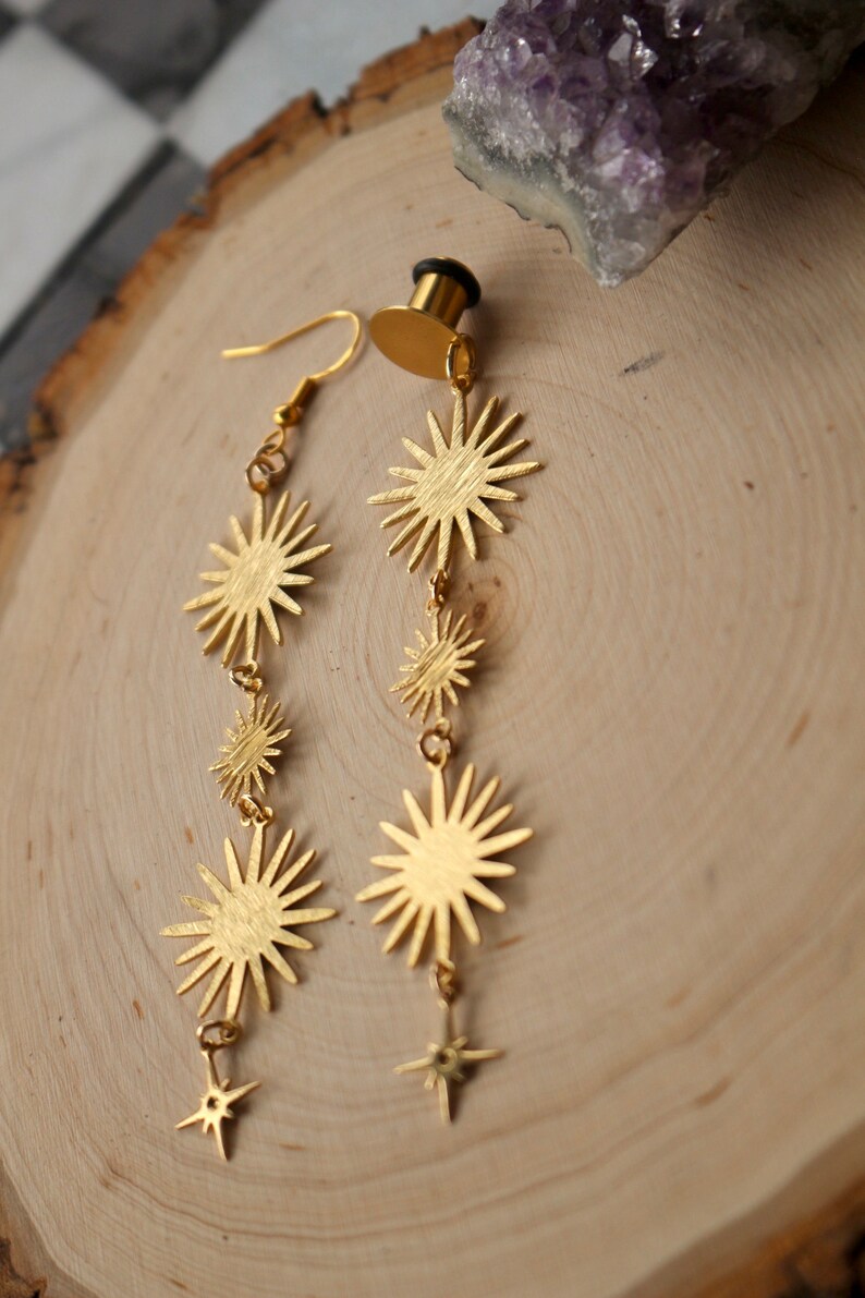 May include: A pair of gold-toned earrings with a dangling star design. Each earring features a series of five star-shaped charms, with the largest star at the top and the smallest star at the bottom.