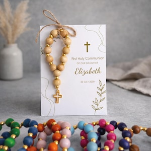 May include: A white rectangular card featuring a gold cross, the text First Holy Communion of our daughter Elizabeth 22 July 2030, and a small wooden bead rosary with a gold cross charm tied to the top with a thin brown twine string.