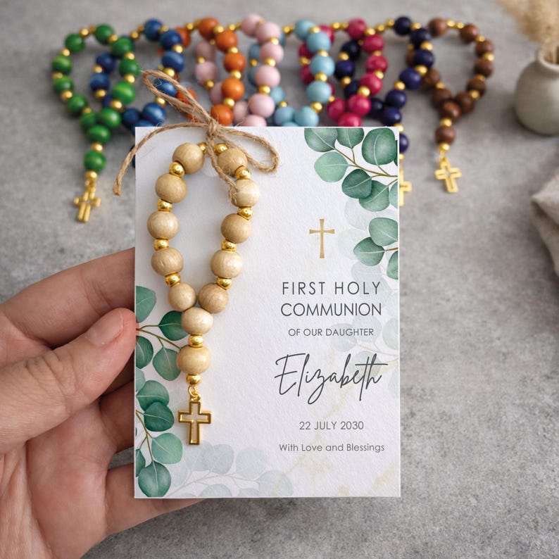 May include: A First Holy Communion card for Elizabeth dated 22 July 2030, featuring green eucalyptus leaves and a gold cross. A small rosary with light wooden beads, gold spacers, and a gold cross is tied to the card with twine.