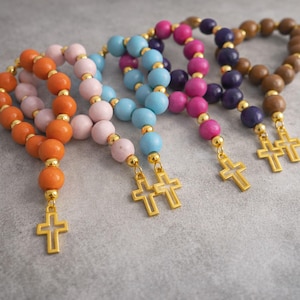 May include: Five rosary bracelets featuring round wooden beads in orange, light pink, light blue, magenta, and brown. Each bracelet is accented with small gold-toned metal spacer beads and a gold-toned metal cross charm dangling from the end.