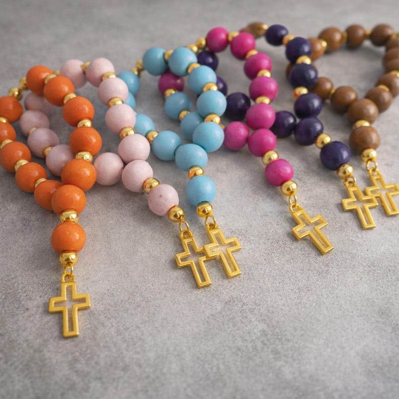 May include: Five rosary bracelets featuring round wooden beads in orange, light pink, light blue, magenta, and brown. Each bracelet is accented with small gold-toned metal spacer beads and a gold-toned metal cross charm dangling from the end.