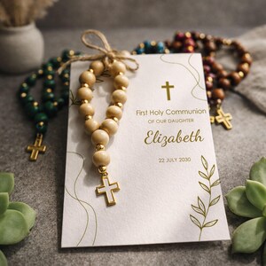 May include: A First Holy Communion card for Elizabeth dated 22 July 2030, featuring a gold cross and leaf illustrations. A small rosary made of light wood beads, gold spacers, and a white cross charm is tied to the card with twine.
