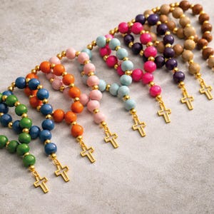 May include: A collection of rosary bracelets featuring round beads in various colors including green, blue, orange, pink, light blue, magenta, purple, and brown. Each bracelet is accented with small gold-tone beads and a gold-tone cross charm.