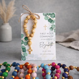 May include: A First Holy Communion card for Elizabeth dated 22 July 2030, featuring green eucalyptus leaves, a gold cross, and a wooden bead rosary tied with twine. Additional colorful wooden bead rosaries are arranged in the foreground.