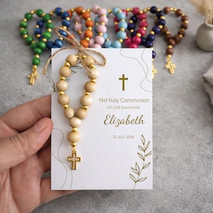 May include: A white card featuring a gold cross, leaf illustrations, and the text First Holy Communion of our daughter Elizabeth 22 July 2030. A small rosary with light wood beads and gold spacers is tied to the card with twine. Other colourful rosaries are visible in the background.