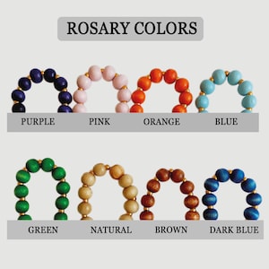 May include: A color chart titled ROSARY COLORS displaying eight circular arrangements of wooden beads separated by small gold-toned spacers. The colors are labeled as purple, pink, orange, blue, green, natural, brown, and dark blue.