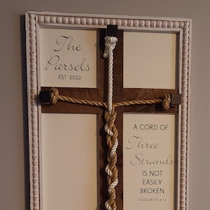 May include: A white wooden frame with a brown wooden cross inside. The cross is decorated with two ropes, one white and one tan, that are braided together. The frame has the text "The Parsels EST 2022" on the top and "A CORD OF Three Strands IS NOT EASILY BROKEN ECCLESIASTES 4 12" on the bottom.