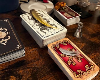 May include: Several tarot decks are arranged on a wooden surface. One deck has a black cover with a crescent moon design. Another deck has a red cover with gold filigree. A yellow feather rests on a deck with a white cover.