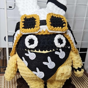 May include: A handmade crochet bunny doll with white ears, a black and white aviator cap, and a yellow body. The bunny wears a black bandana with white rabbit silhouettes and gold-trimmed goggles.