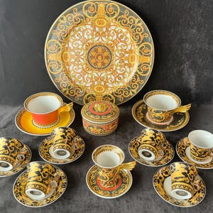 May include: A luxury porcelain tea set featuring a large decorative plate, a small round trinket box, saucers, and teacups. The set has a black, gold, and white color scheme with ornate patterns and designs. The teacups have a unique wing-shaped handle.
