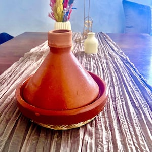 May include: A terracotta tagine with a round base and a conical lid. The tagine is sitting on a light brown table runner.
