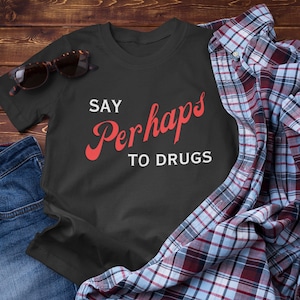 Puede incluir: Camiseta negra con texto blanco que dice "Say Perhaps to Drugs" en rojo, en letra cursiva.