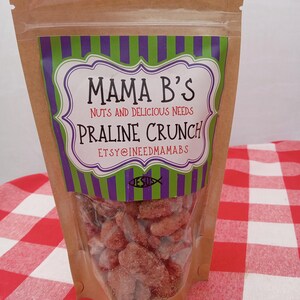 May include: A clear plastic bag filled with praline crunch candy. The bag has a brown paper exterior and a label with the text "Mama B's Nuts and Delicious Needs Praline Crunch". The label has a purple and green striped border and white text.