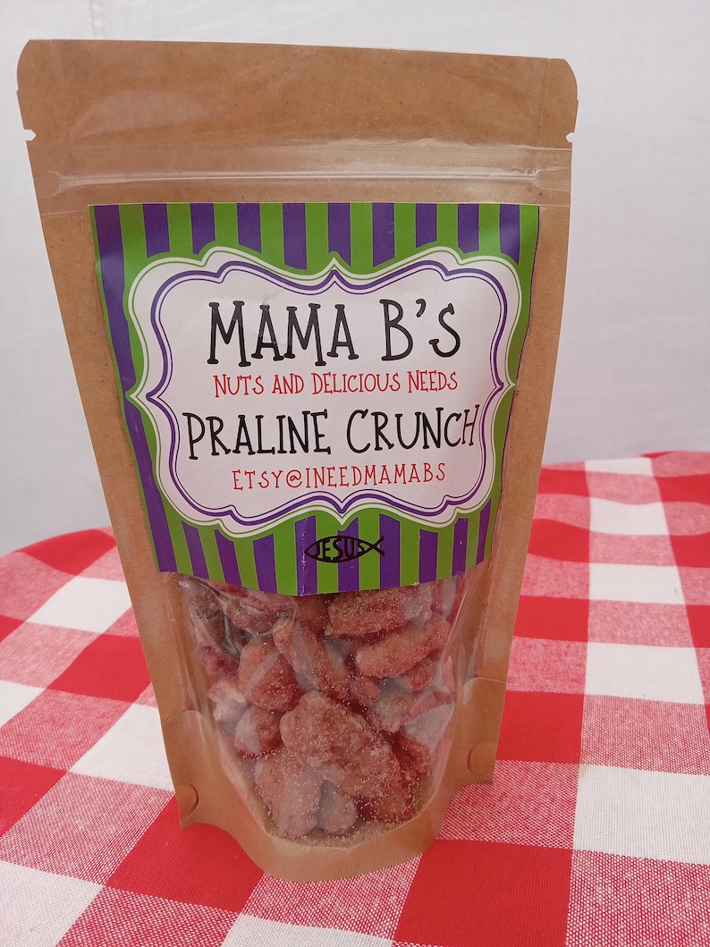 May include: A clear plastic bag filled with praline crunch candy. The bag has a brown paper exterior and a label with the text "Mama B's Nuts and Delicious Needs Praline Crunch". The label has a purple and green striped border and white text.