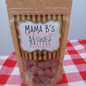 May include: A clear plastic bag filled with brownie brittle. The bag has a brown paper top with the text "Mama B's" and "Brownie Brittle". The bag is on a red and white checkered tablecloth.