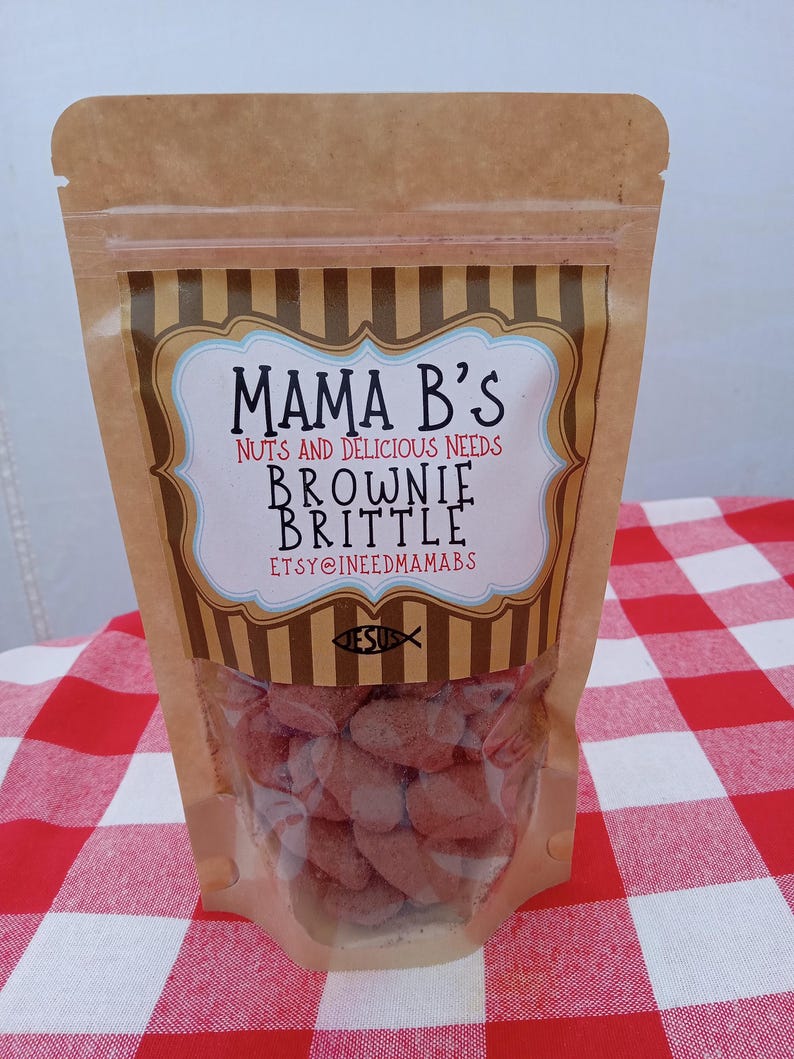May include: A clear plastic bag filled with brownie brittle. The bag has a brown paper top with the text "Mama B's" and "Brownie Brittle". The bag is on a red and white checkered tablecloth.