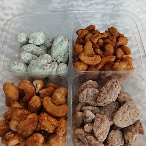 May include: A clear plastic container divided into four sections, each filled with different types of nuts. The nuts include sugared cashews, sugared pecans, and nuts coated in a white and green candy shell. The nuts are arranged on a white surface.