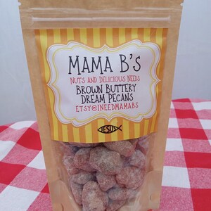 May include: A clear plastic bag filled with brown sugared pecans. The bag has a brown paper top with a yellow and white label that reads "Mama B's" and "Brown Buttery Dream Pecans". The background is a red and white checkered tablecloth.