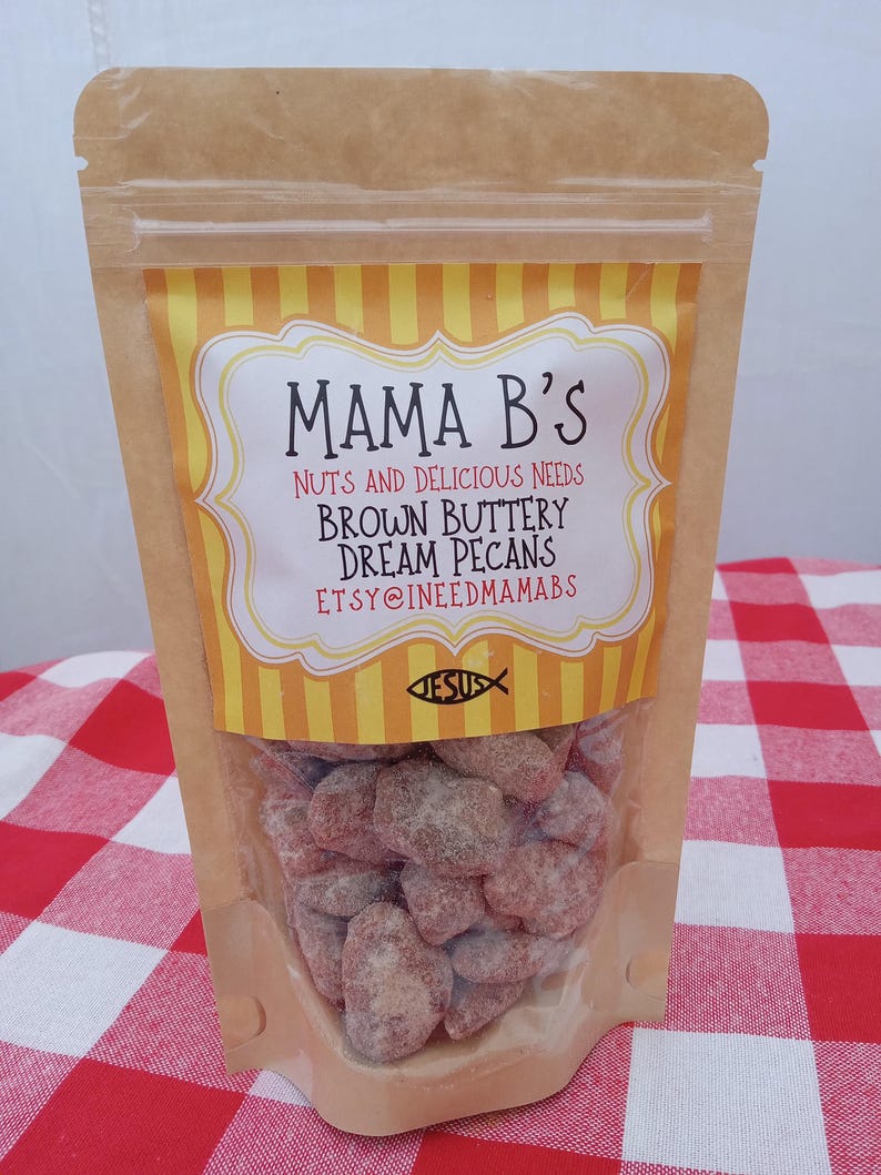 May include: A clear plastic bag filled with brown sugared pecans. The bag has a brown paper top with a yellow and white label that reads "Mama B's" and "Brown Buttery Dream Pecans". The background is a red and white checkered tablecloth.