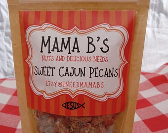 Puede incluir: Una bolsa de papel marrón con una ventana de plástico transparente, que contiene nueces de pecán cajún dulces. La bolsa tiene una etiqueta a rayas rojas y blancas con el texto "Mama B's Nuts and Delicious Needs Sweet Cajun Pecans Etsy@ineedmamabs".