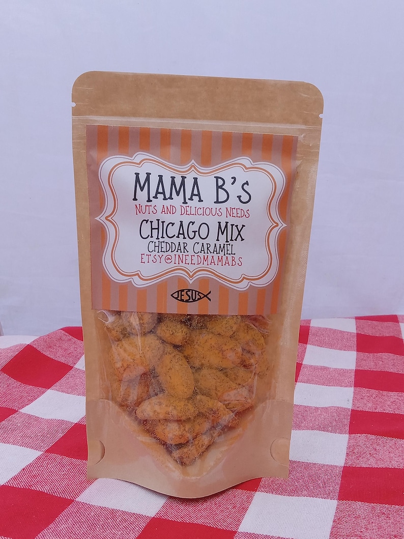 May include: A clear plastic bag filled with Chicago Mix cheddar caramel snacks. The bag has a brown paper exterior with a label that reads "Mama B's Nuts and Delicious Needs Chicago Mix Cheddar Caramel". The bag is on a red and white checkered surface.