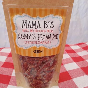 May include: A clear plastic bag filled with pecan pie pieces. The bag has a brown paper exterior with a label that reads "Mama B's Nanny's Pecan Pie." The label has a blue and white border with an orange and white striped background. The bag is on a red and white checkered surface.