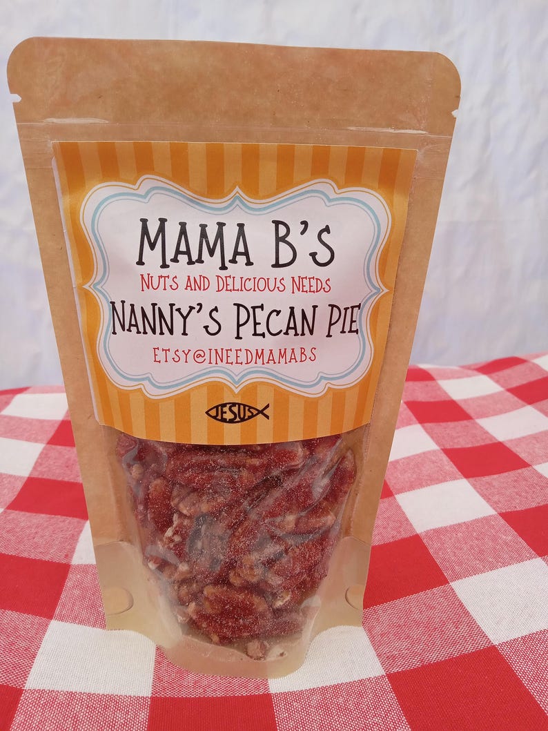 May include: A clear plastic bag filled with pecan pie pieces. The bag has a brown paper exterior with a label that reads "Mama B's Nanny's Pecan Pie." The label has a blue and white border with an orange and white striped background. The bag is on a red and white checkered surface.