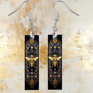 May include: A pair of silver earrings with black rectangular pendants featuring a gold bee design and intricate floral patterns.