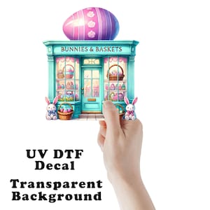 May include: A colorful UV DTF decal featuring a whimsical Easter shop facade with the text "Bunnies & Baskets." The shop is adorned with Easter eggs and baskets. A large, striped egg sits atop the building. The decal has a transparent background.