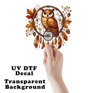 May include: A UV DTF decal with a transparent background. The design features a detailed owl perched within a dreamcatcher, surrounded by autumn leaves and feathers. The owl has brown and orange feathers.