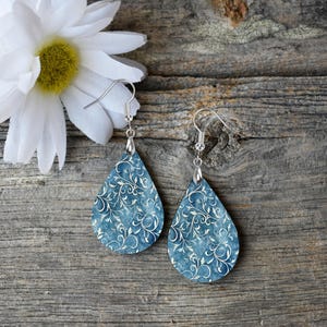 Pu&ograve; includere: Un paio di orecchini a forma di goccia con un motivo floreale blu e bianco. Gli orecchini sono in legno e hanno ganci d'argento.