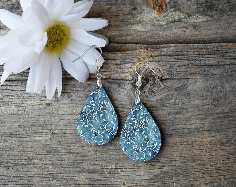 Pu&ograve; includere: Un paio di orecchini a forma di goccia con un motivo floreale blu e bianco. Gli orecchini sono in legno e hanno ganci d'argento.