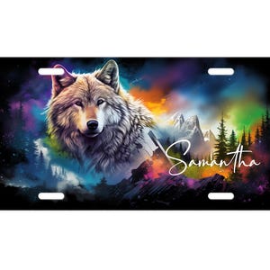 May include: License plate featuring a detailed wolf illustration with a colorful, abstract background. The design includes mountains, trees, and the name "Samantha" in a script font. The plate has four mounting holes.