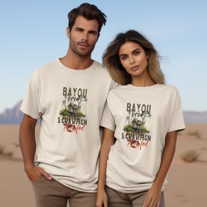 May include: Two beige t-shirts with a graphic print of a house on stilts over water with the text "Bayou bread & crawfish fed".