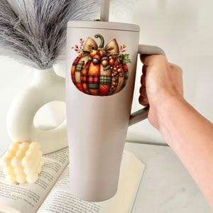 May include: A gray stainless steel tumbler with a handle and a straw. The tumbler features a colorful illustration of a plaid pumpkin with a bow and fall foliage.