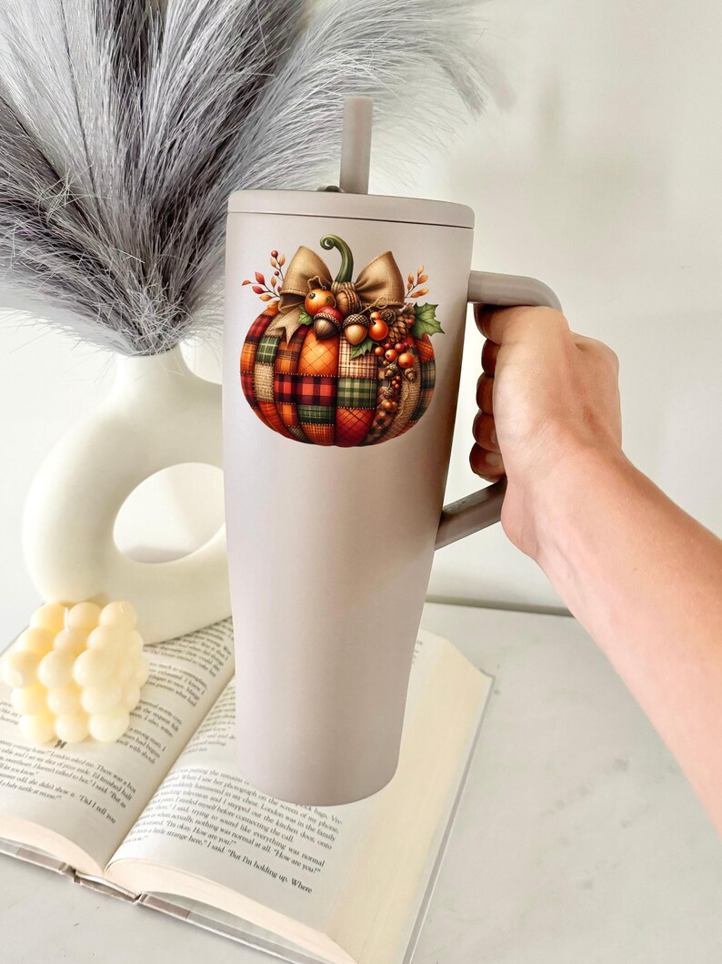 May include: A gray stainless steel tumbler with a handle and a straw. The tumbler features a colorful illustration of a plaid pumpkin with a bow and fall foliage.