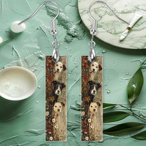 May include: A pair of silver dangle earrings with rectangular charms featuring a whimsical illustration of three dogs with floral accents. The charms are a light brown color with a white background.