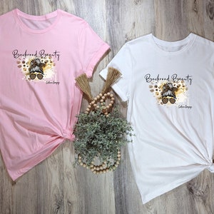 May include: Two t-shirts with a graphic design of a woman with a messy bun and sunglasses. The text "Backroad Beauty" is above the graphic. The t-shirts are pink and white.