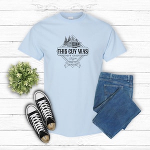 May include: Light blue t-shirt with a black and white graphic design of a cabin in the woods. The graphic says "This Guy Was Off Grid Before It Was Cool."
