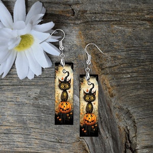 May include: A pair of dangle earrings featuring a black cat sitting on a jack-o'-lantern. The cat is wearing a witch's hat and has a mischievous expression. The earrings are set against a dark background with orange and yellow accents.