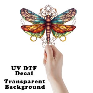 May include: A colorful dragonfly decal with a transparent background. The dragonfly features intricate wing designs in shades of blue, orange, and yellow. The text "UV DTF Decal" and "Transparent Background" are displayed below the dragonfly.