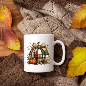 May include: White ceramic mug featuring a fall-themed design with a plaid pumpkin, bow, and small pumpkins. The mug is set on a brown knitted surface with colorful autumn leaves.