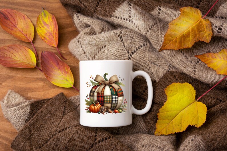 May include: White ceramic mug featuring a fall-themed design with a plaid pumpkin, bow, and small pumpkins. The mug is set on a brown knitted surface with colorful autumn leaves.
