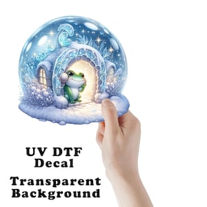 May include: A UV DTF decal featuring a snow globe illustration with a green frog sitting inside a glowing, snow-covered igloo. The scene includes blue-toned winter foliage, sparkling stars, and a soft light emanating from the igloo entrance. The text UV DTF Decal Transparent Background is printed below the graphic.