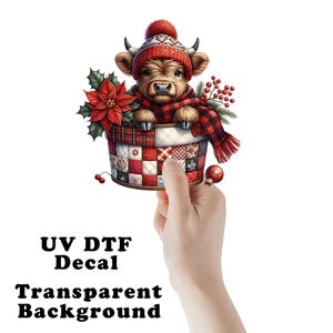 May include: A UV DTF decal with a transparent background. The image features a brown Highland cow wearing a red and white knit hat and scarf, peeking out of a patchwork basket. A poinsettia and berries add festive detail.