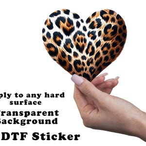 May include: A heart-shaped sticker with a leopard print design, held by a hand. The sticker has a transparent background and is designed to apply to any hard surface. The text on the image reads: "Apply to any hard surface, Transparent Background, UV DTF Sticker."