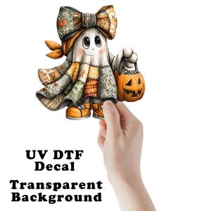 May include: A UV DTF decal featuring a cartoon ghost character wearing a large patchwork-patterned bow and a matching patchwork-style cloak. The ghost has large eyes, a small smile, and is holding an orange jack-o'-lantern bucket with a black handle.